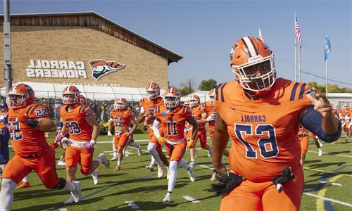 Carroll University football players in bright orange uniforms run onto the field, led by a player wearing number 67. 